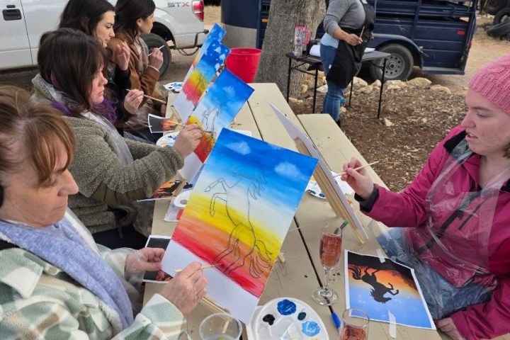 People outdoors painting landscapes at picnic tables, guided by an instructor.