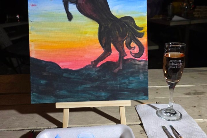 Painting of a rearing horse at sunset with paint palette and wine glass on table.