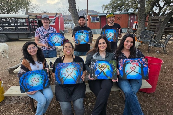 Group of seven people display heart-themed paintings outdoors on a farm.