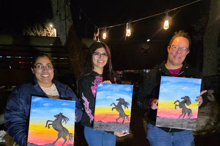 Three people hold paintings of rearing horses at a nighttime art event.