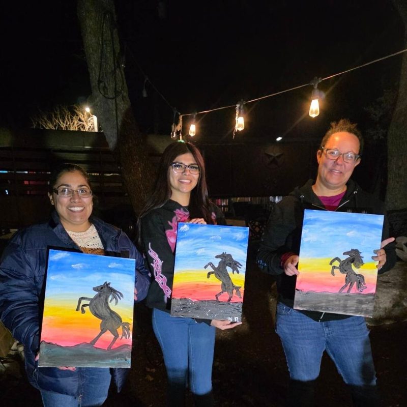 Three people hold paintings of rearing horses at a nighttime art event.