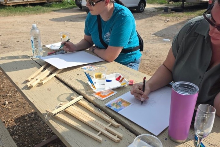 Two people painting outdoors at a picnic table with art supplies.
