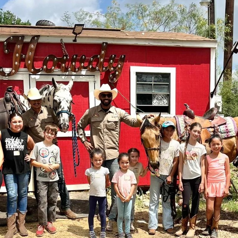 People and two horses in front of a red building with 'Howdy' sign.