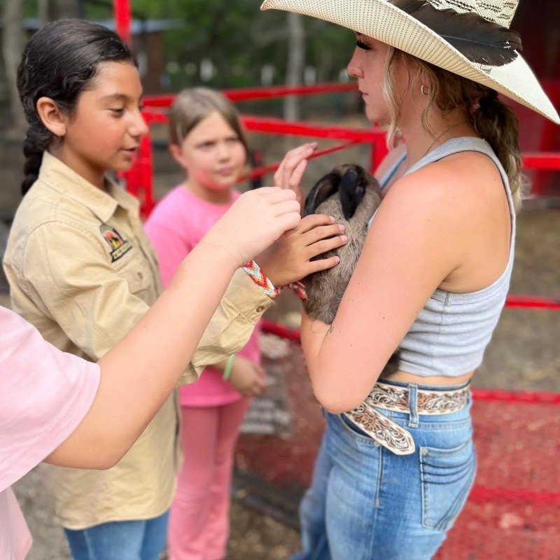 Children petting an animal held by a girl in a cowboy hat and jeans.